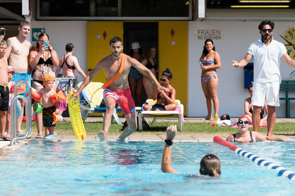 Sanitäter springt vom Beckenrand ins Wasser, um eine Person in Not zu retten; rundherum stehen Menschen, die zusehen, Wasserrettung, Übung,